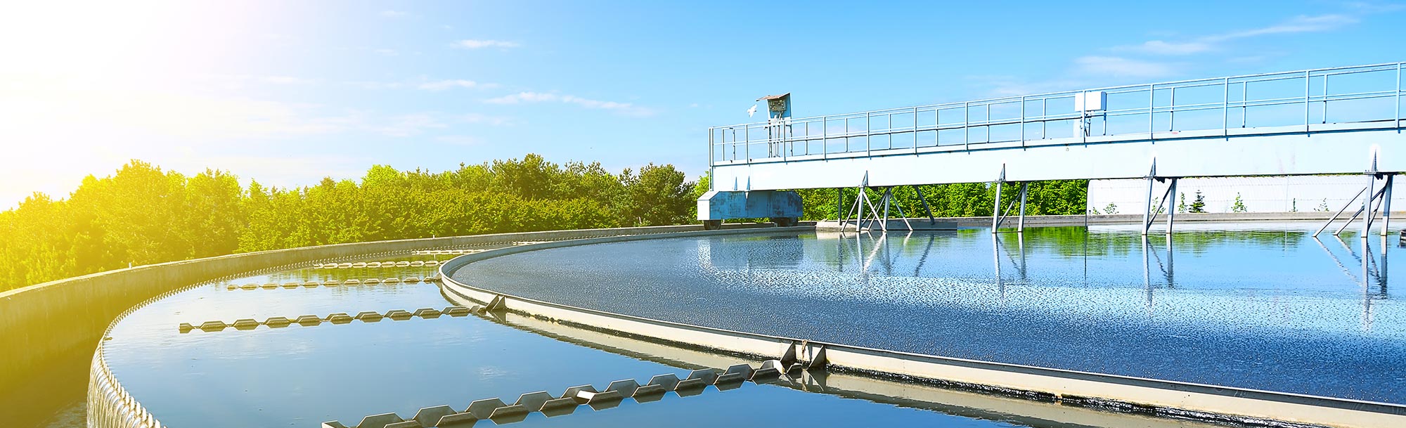Kläranlage mit rundem Nachklärbecken und Steg über der Wasseroberfläche bei Sonnenschein.