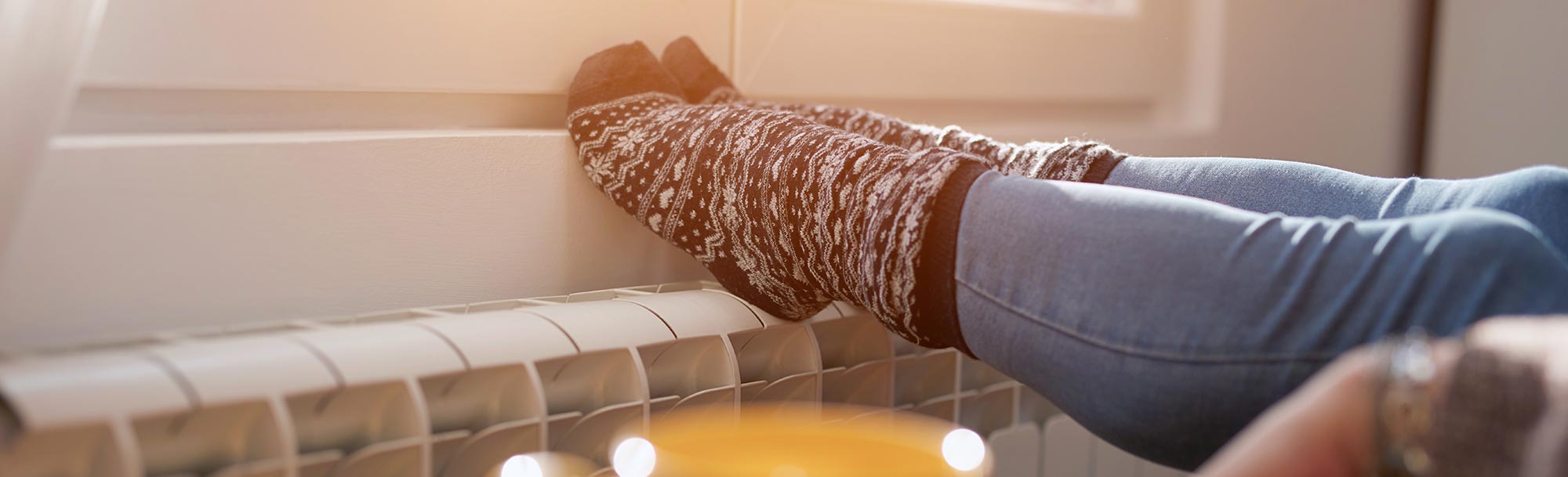 Person mit warmen Wollsocken legt die Füße auf einen Heizkörper, im Vordergrund steht eine Tasse.