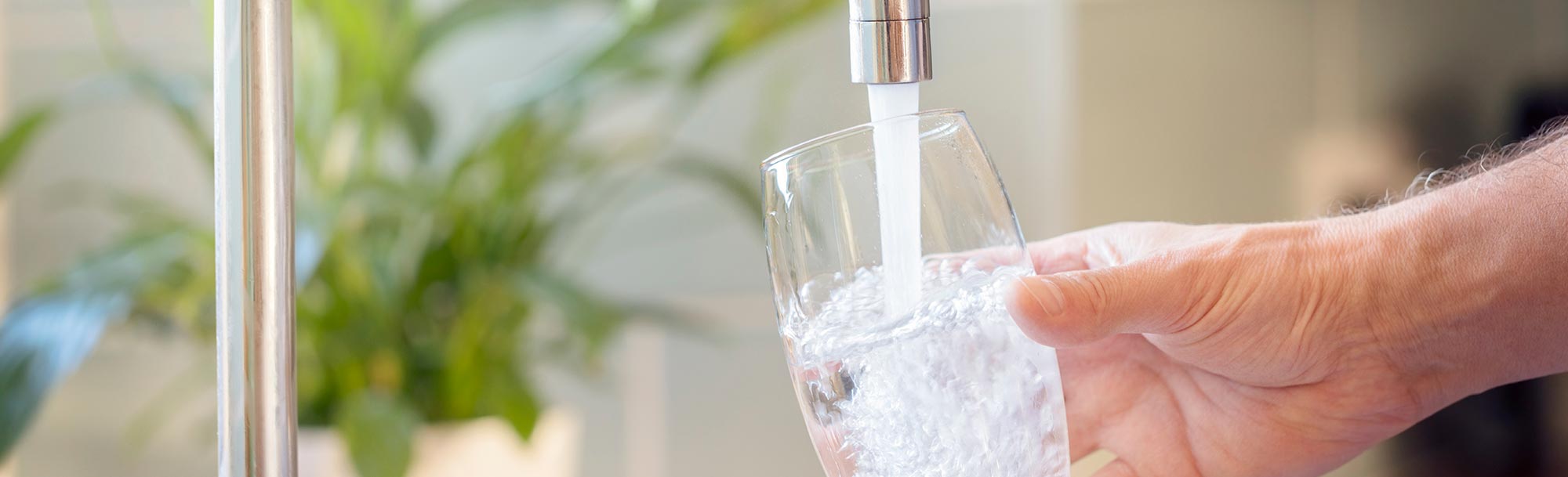 Hand hält ein Glas unter einen laufenden Wasserhahn, das mit klarem Trinkwasser gefüllt wird.