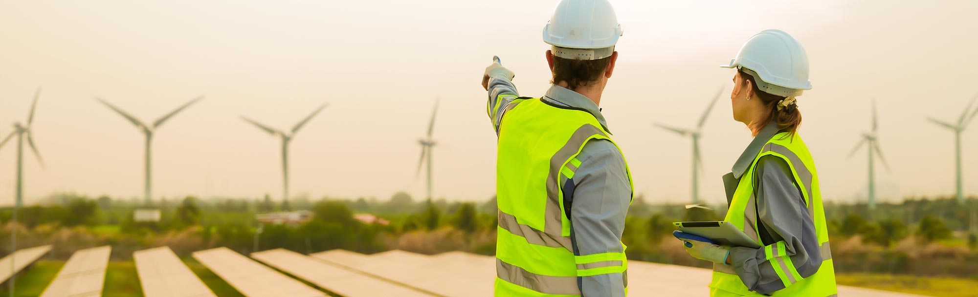 Zwei Ingenieure in Warnwesten und Schutzhelmen betrachten eine Anlage mit Windkrafträdern und Solarpaneelen; symbolisiert technische Fachkompetenz und nachhaltige Energieerzeugung.
